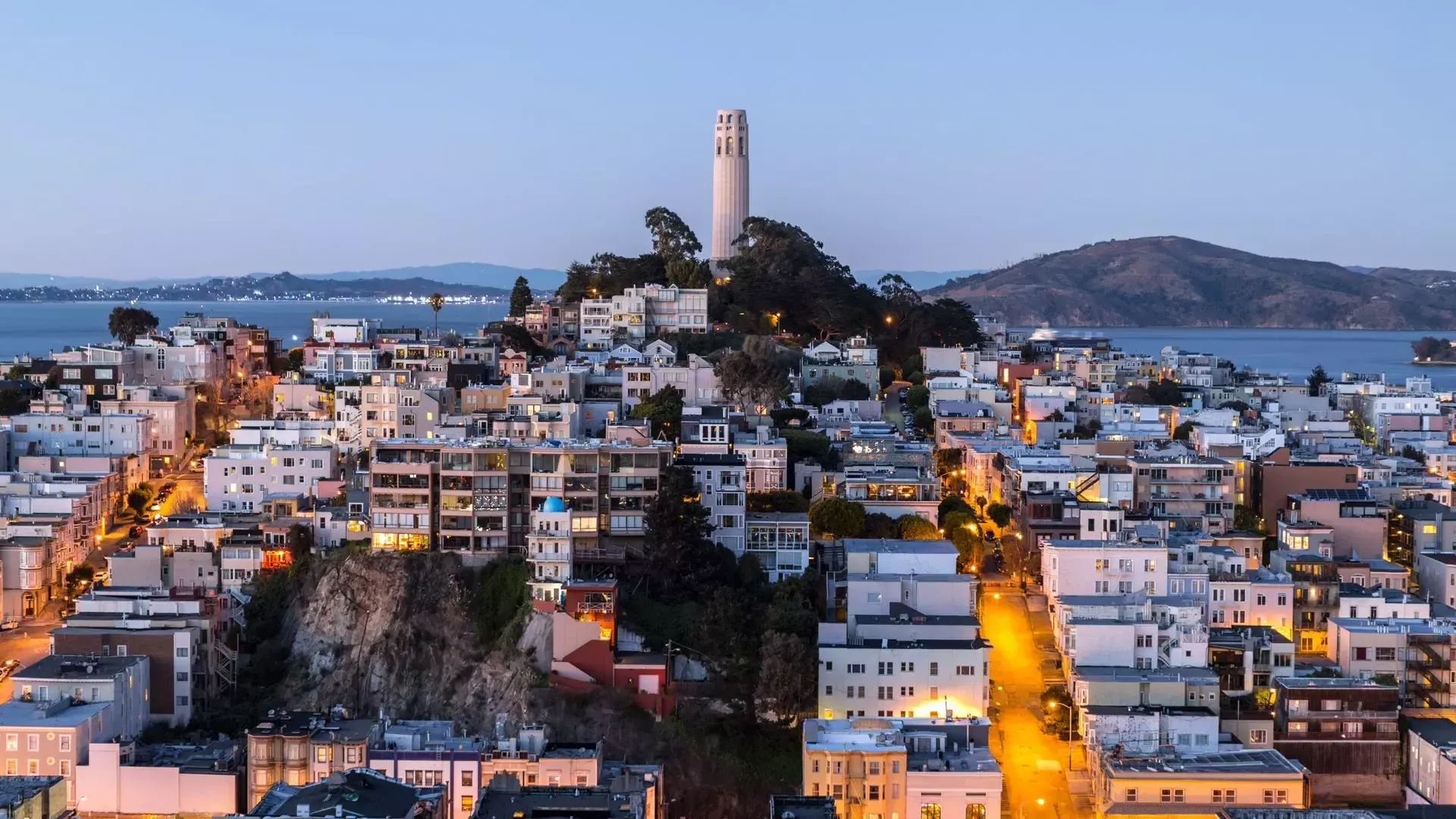 Coit Tower em São Francisco, ao anoitecer, com ruas iluminadas à sua frente e a Baía de São Francisco atrás.